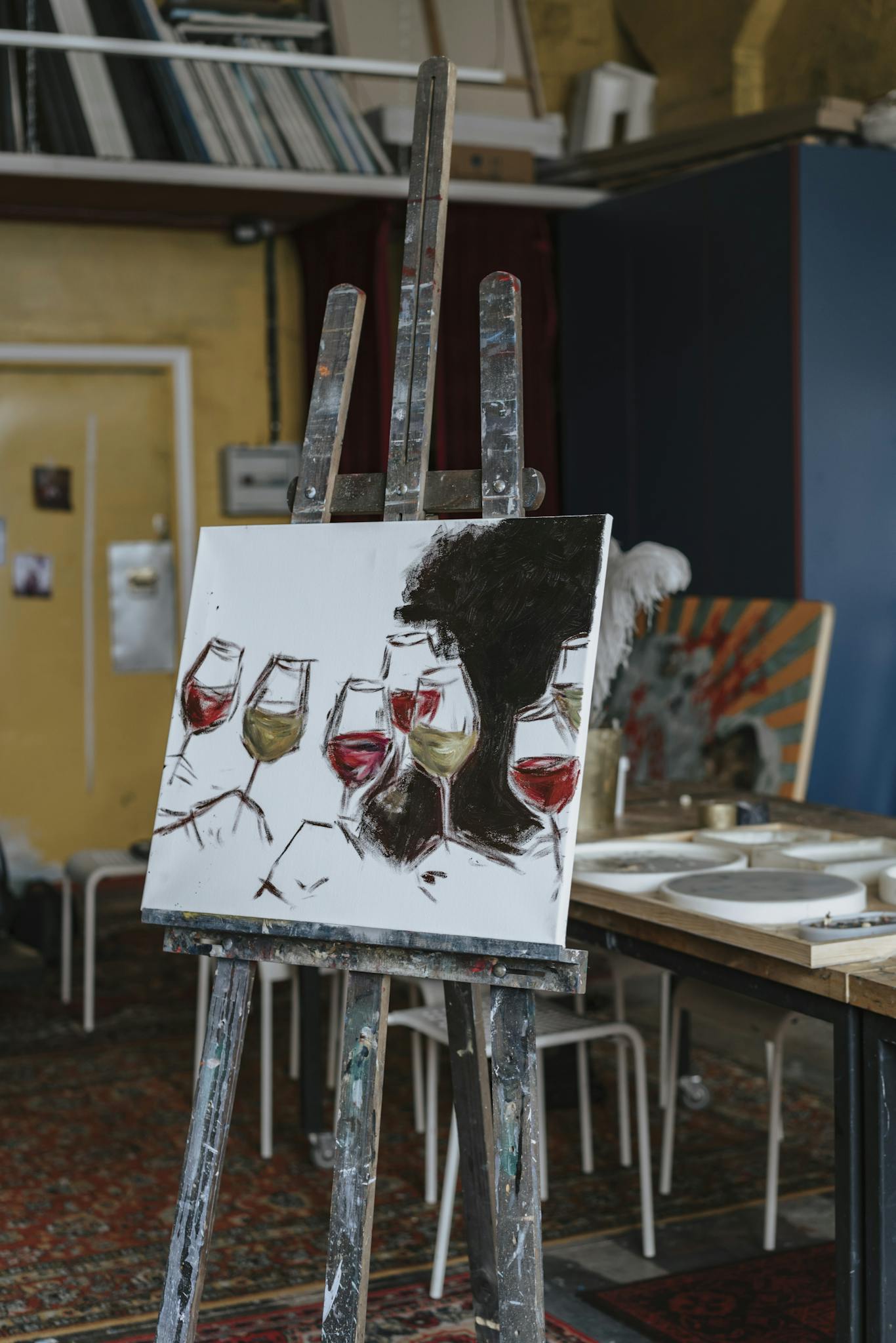 A canvas with abstract wine glass painting on an easel in a cluttered art studio.